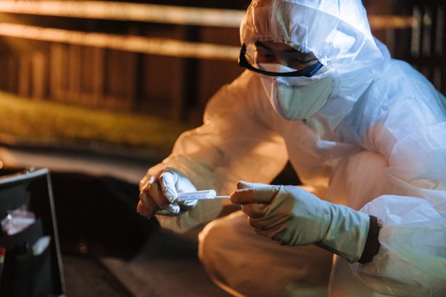 A forensic investigator in protective gear collects evidence outdoors at night.